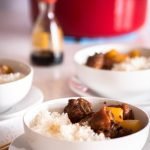A close-up shot of a bowl filled with tender sweet and sour spare ribs, coated in a rich, flavorful sauce, and paired with bright yellow pineapple chunks. The dish is served over steamed white rice in a white ceramic bowl. In the lower portion of the image, a second bowl of the same dish is placed on a white plate with chopsticks resting beside it. A bold red text overlay reads: 'Delicious Sweet & Sour Spare Ribs Recipe.