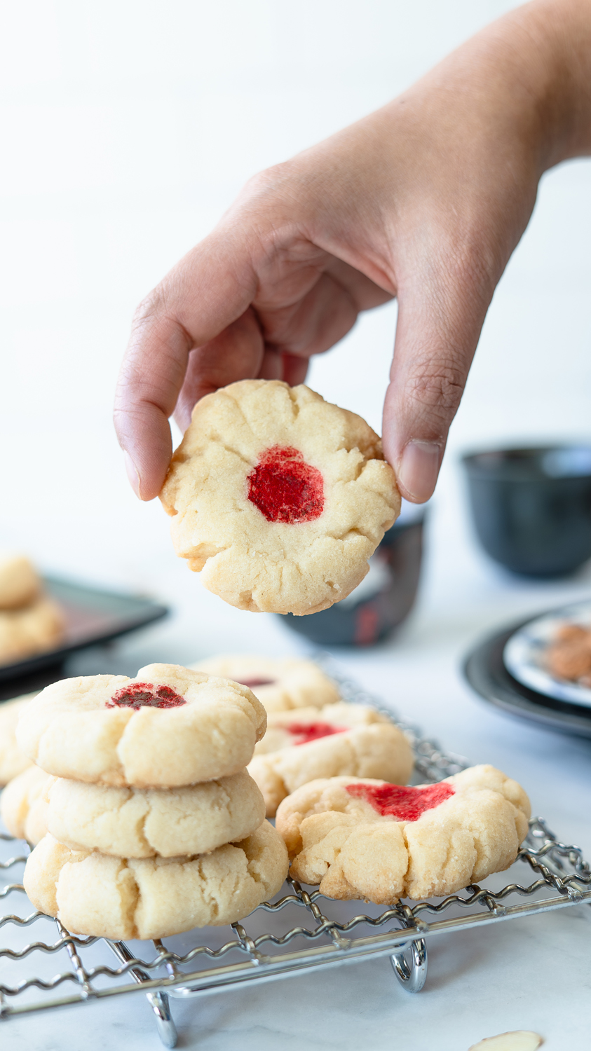 Hand holding a crumbly Chinese almond cookie with a bright red dot above a stack of red dot cookies on a cooling rack.