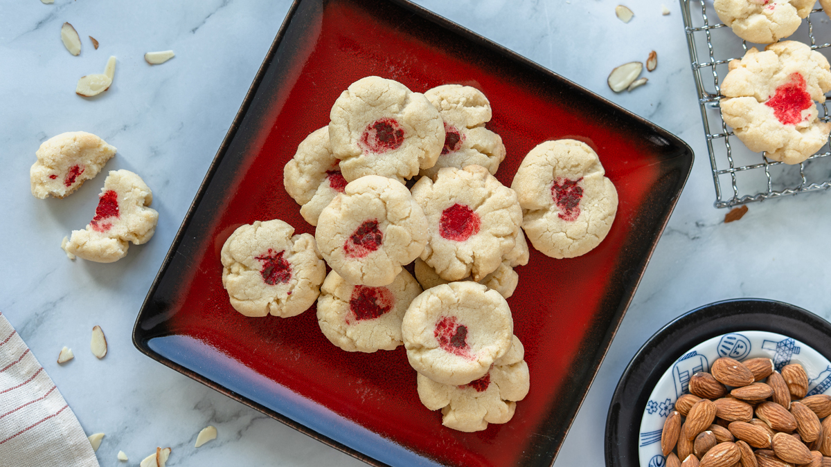 Overhead view of crumbly Chinese almond cookies with red dots piled on a red square plate, with sliced almonds and a bowl of whole almonds nearby.