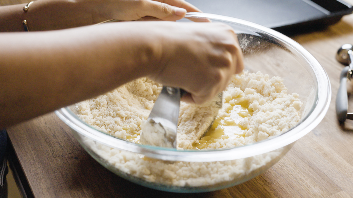 Pastry blender mixing egg into the crumbly flour mixture in a glass bowl.