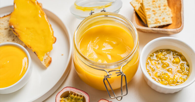 A glass jar of golden lilikoi butter surrounded by a slice of toast spread with lilikoi butter, a small bowl of lilikoi butter, fresh halved passion fruit, crackers on a wooden board, and a small bowl of passion fruit pulp, all on a white surface.