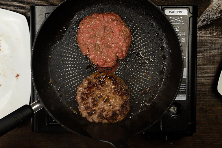 Two loco moco beef patties cooking in a skillet — one raw side up and one seared brown side showing the crust from pan frying