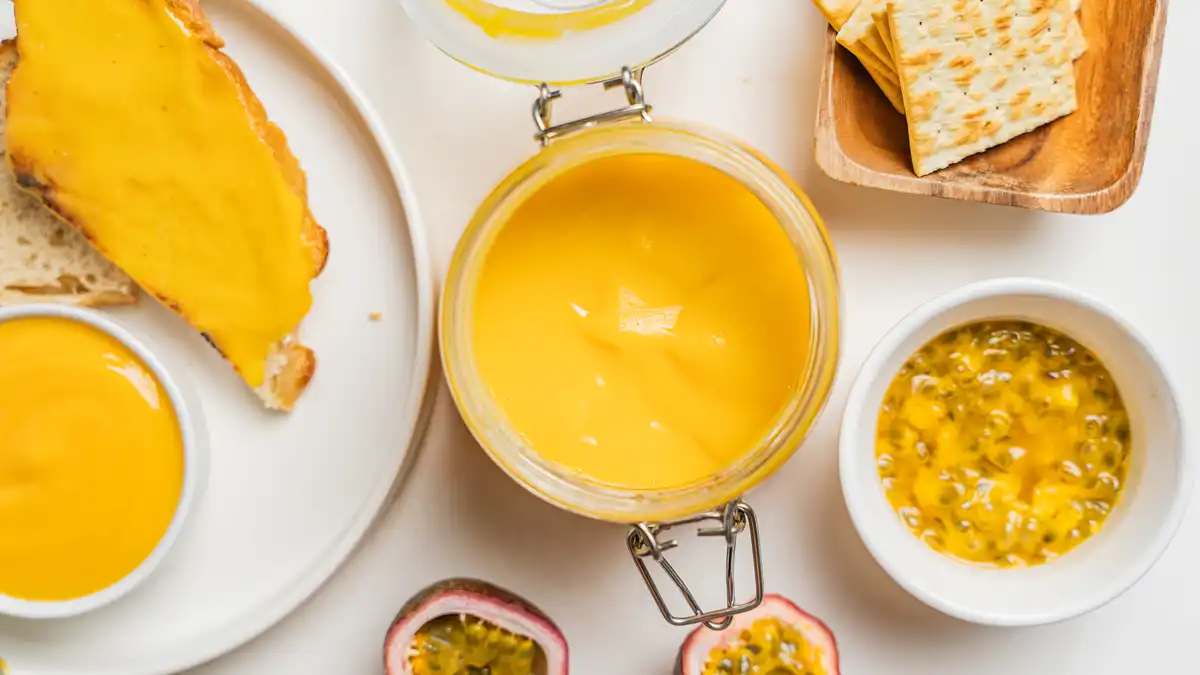 Overhead shot of lilikoi butter in an open glass jar surrounded by toast spread with lilikoi butter, a small bowl of lilikoi butter, a bowl of fresh lilikoi pulp, halved passion fruit, and crackers on a wooden tray, all on a white surface