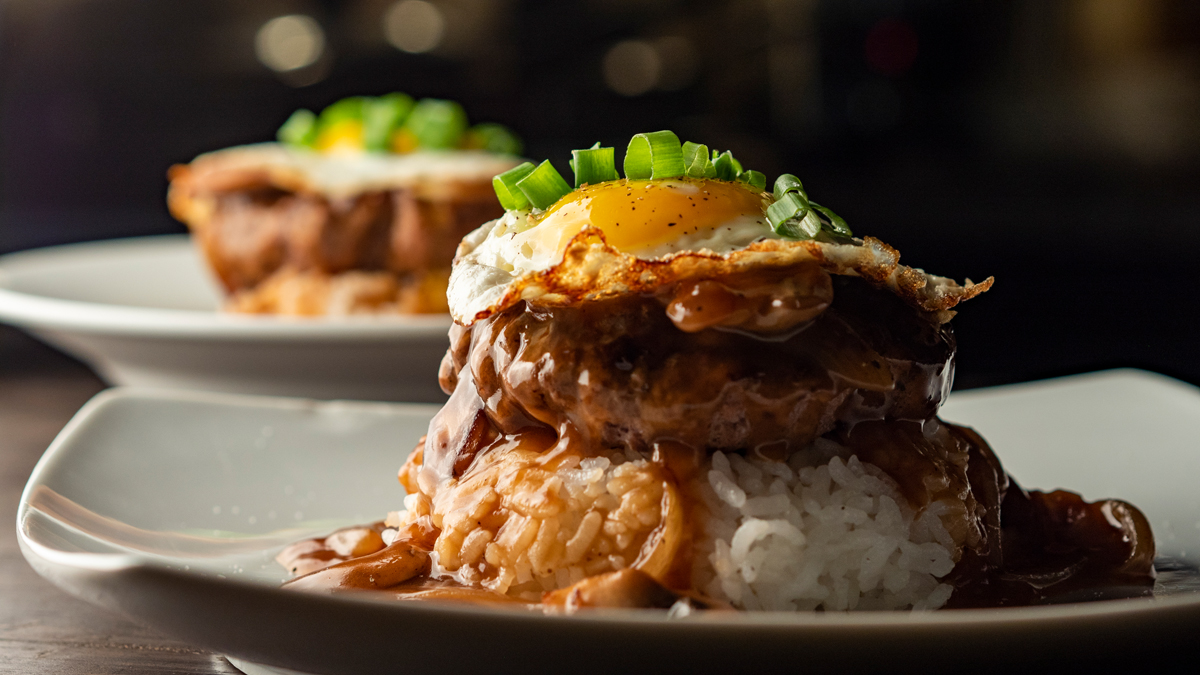 Close-up of a loco moco recipe showing a thick beef patty smothered in glossy brown gravy over white rice, topped with a crispy-edged fried egg and fresh green onions, with a second serving blurred in the background