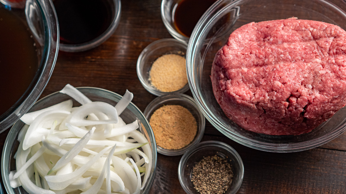 Loco moco recipe ingredients in glass bowls on a dark wood surface — ground beef, sliced sweet onion, garlic powder, seasoning salt, black pepper, shoyu, and Worcestershire sauce