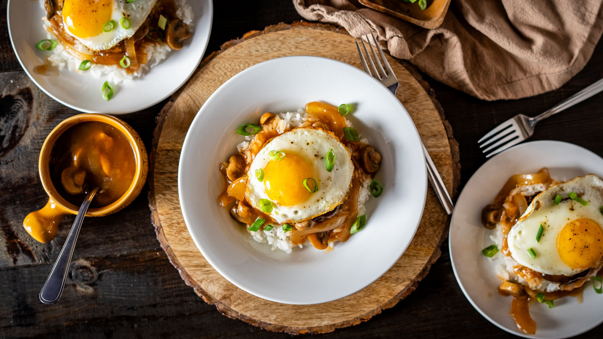 Three servings of loco moco recipe overhead — white rice topped with beef patty, mushroom brown gravy, and sunny-side up eggs garnished with green onions, with a small ramekin of extra gravy on the side
