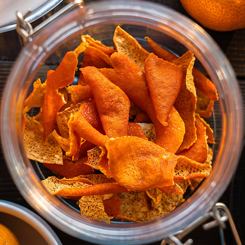 Close-up overhead view of dried orange peel chips piled inside an open glass clamp jar, showing the vivid orange color and textured surface of the dried peels