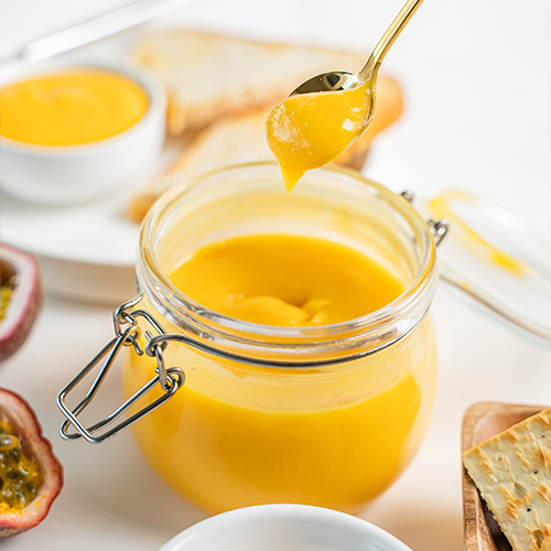 A gold spoon lifting a drizzle of smooth, glossy lilikoi butter from an open glass jar, with halved passion fruit, toast, crackers, and a small bowl of lilikoi butter in the background