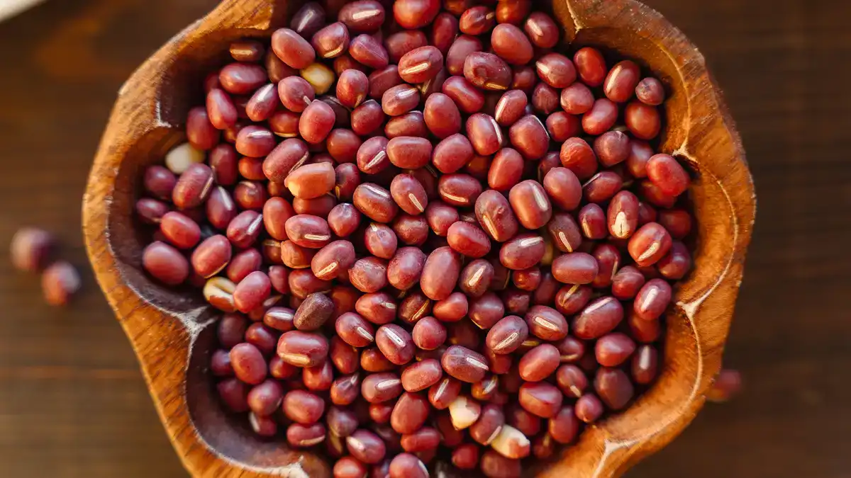 A close-up overhead shot of dried azuki beans filling a carved wooden bowl, set on a dark wood surface with a few scattered beans alongside.