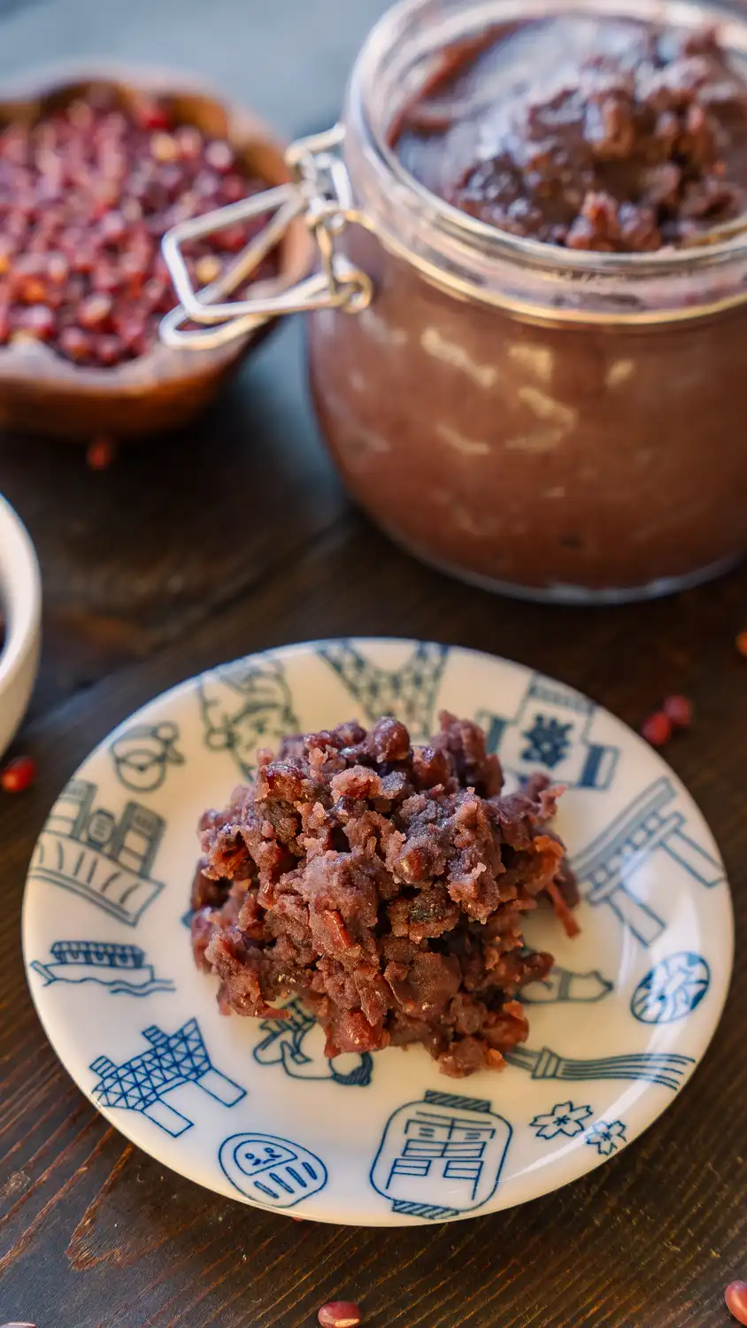 Homemade tsubuan anko piled on a decorative blue and white plate, with a large glass jar of red bean paste and a wooden bowl of dried azuki beans in the background on a dark wood surface.