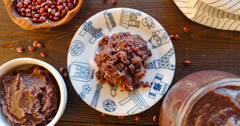 Two styles of homemade anko on a dark wood surface โ chunky tsubuan on a decorative plate and smooth koshian in a ceramic bowl โ with a jar of red bean paste and a bowl of dried azuki beans in the background.