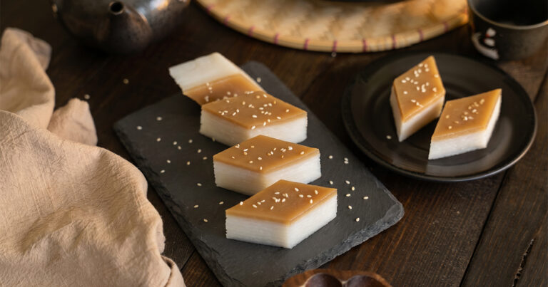 Diamond-cut pieces of layered rice cake on a dark slate board, showing distinct white layers topped with a golden brown sugar layer and scattered sesame seeds, styled with a ceramic teapot, dark plates, and linen cloth on a dark wood table