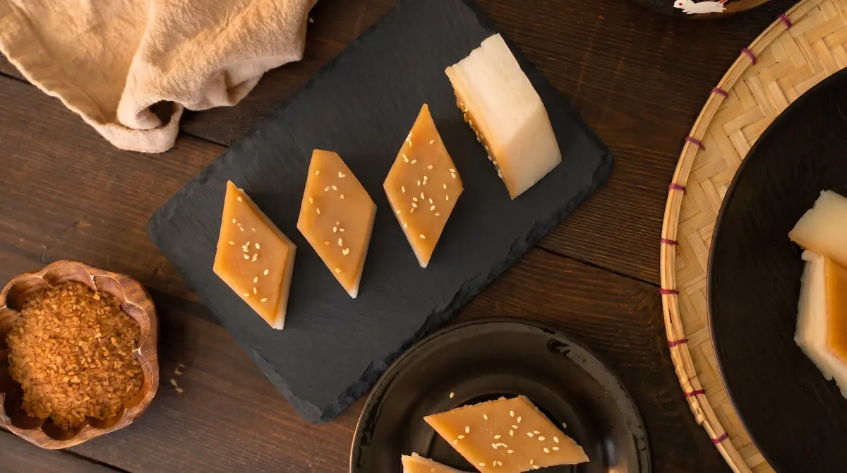 Overhead view of diamond-cut layered rice cake pieces on a dark slate board, brown sugar top layer facing up with sesame seeds, surrounded by a wooden bowl of raw cane sugar, a bamboo steamer, a dark ceramic plate, and linen cloth on a dark wood table