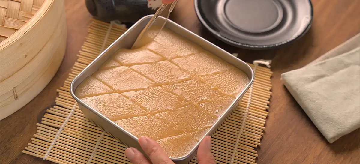 Overhead view of the brown sugar top layer batter being poured from a blue and white ceramic bowl into the square pan over the steamed white layers of layered rice cake inside a bamboo steamer