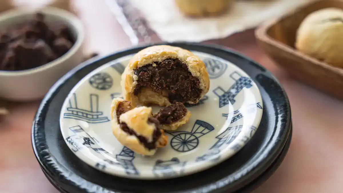A freshly baked Hawaiian manju broken open on a blue and white Japanese-style plate, revealing a rich chocolate anko filling inside the golden flaky pastry crust