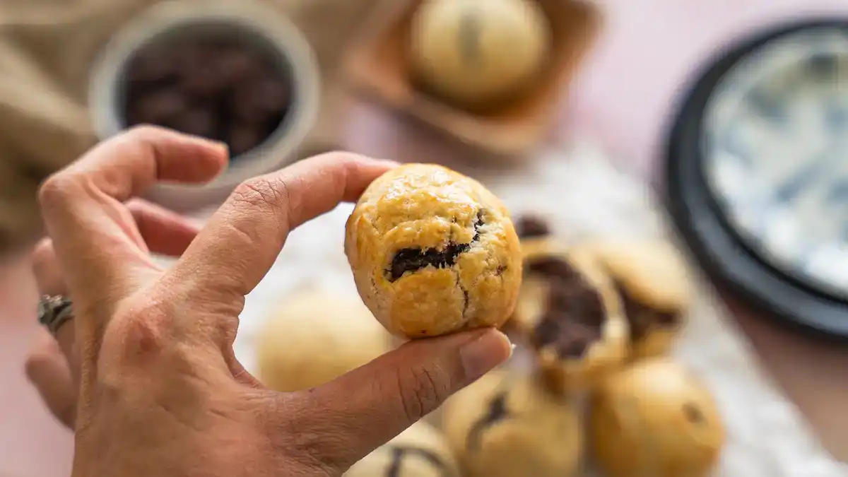 A hand holding a golden baked Hawaiian manju pastry with the anko filling peeking through the flaky buttery crust, with more manju and a bowl of anko in the background