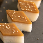 Diamond-cut pieces of layered rice cake on a dark slate board, showing distinct white layers topped with a golden brown sugar layer and scattered sesame seeds, styled with a ceramic teapot, dark plates, and linen cloth on a dark wood table