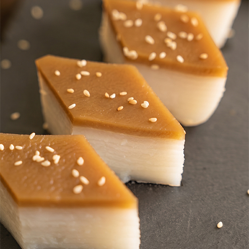 Diamond-cut pieces of layered rice cake on a dark slate board, showing distinct white layers topped with a golden brown sugar layer and scattered sesame seeds, styled with a ceramic teapot, dark plates, and linen cloth on a dark wood table