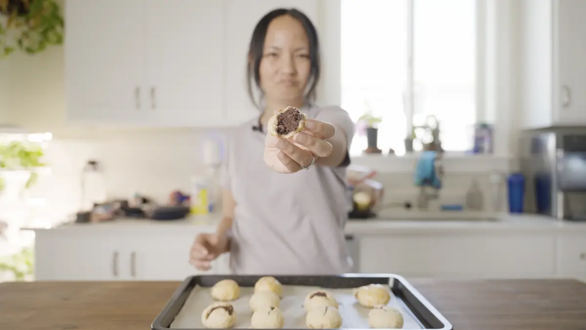 Tani from Ono Hawaiian Recipes holding up a freshly baked Hawaiian manju broken open to show the anko filling, with a full tray of homemade manju on the kitchen counter in front of her