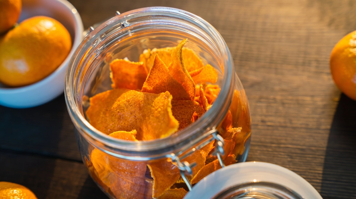 Angled close-up of dried orange peel chips inside an open glass clamp jar with a white ceramic bowl of fresh tangerines in soft warm background light