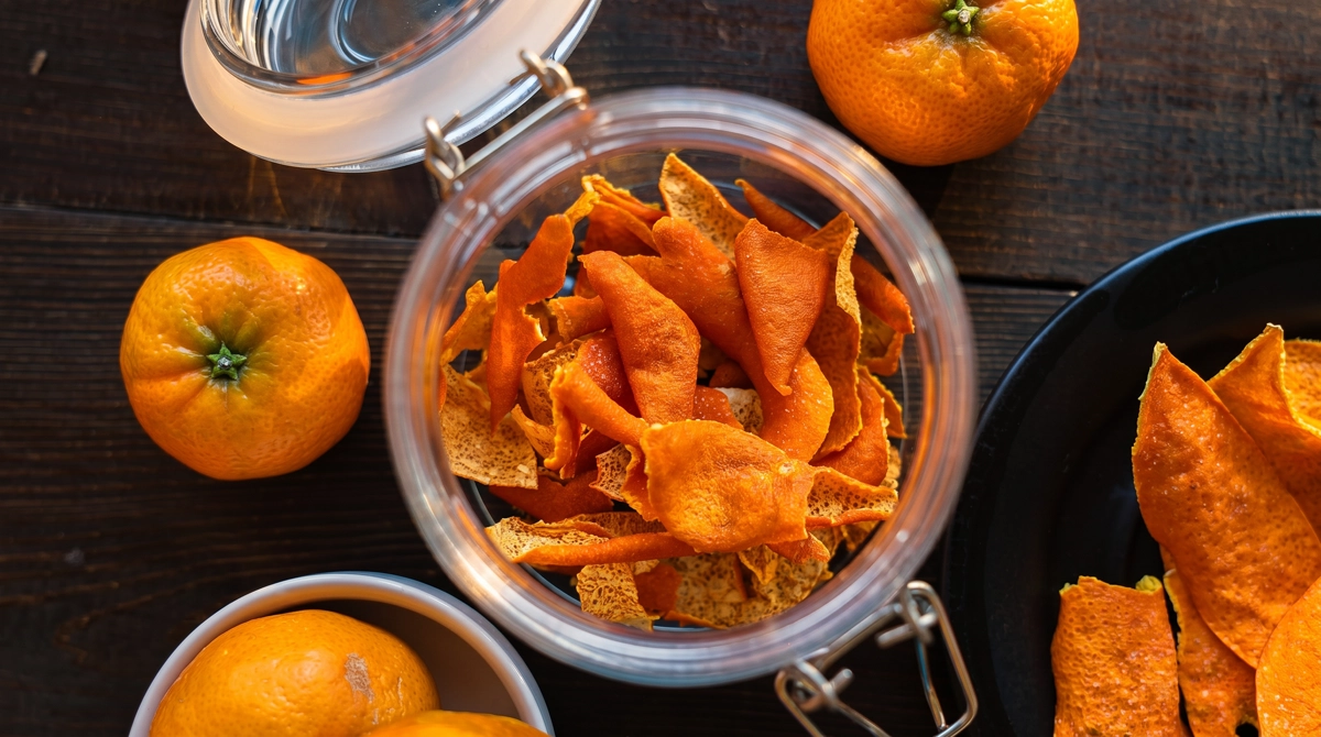 Overhead view of dried orange peel chips in an open glass clamp jar with fresh tangerines and a dark plate of loose dried peels arranged on a dark wood surface in warm natural light