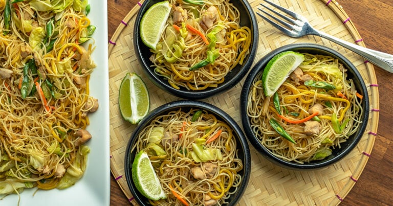 Three black bowls of easy pancit bihon with lime wedges on a woven tray beside a plate of noodles