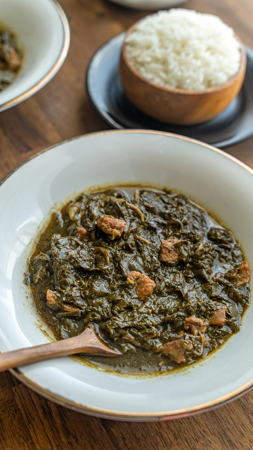 close-up of luau stew in a white bowl with a wooden spoon, dark taro leaves and tender pork pieces in a rich broth, more bowls and steamed rice blurred in the background