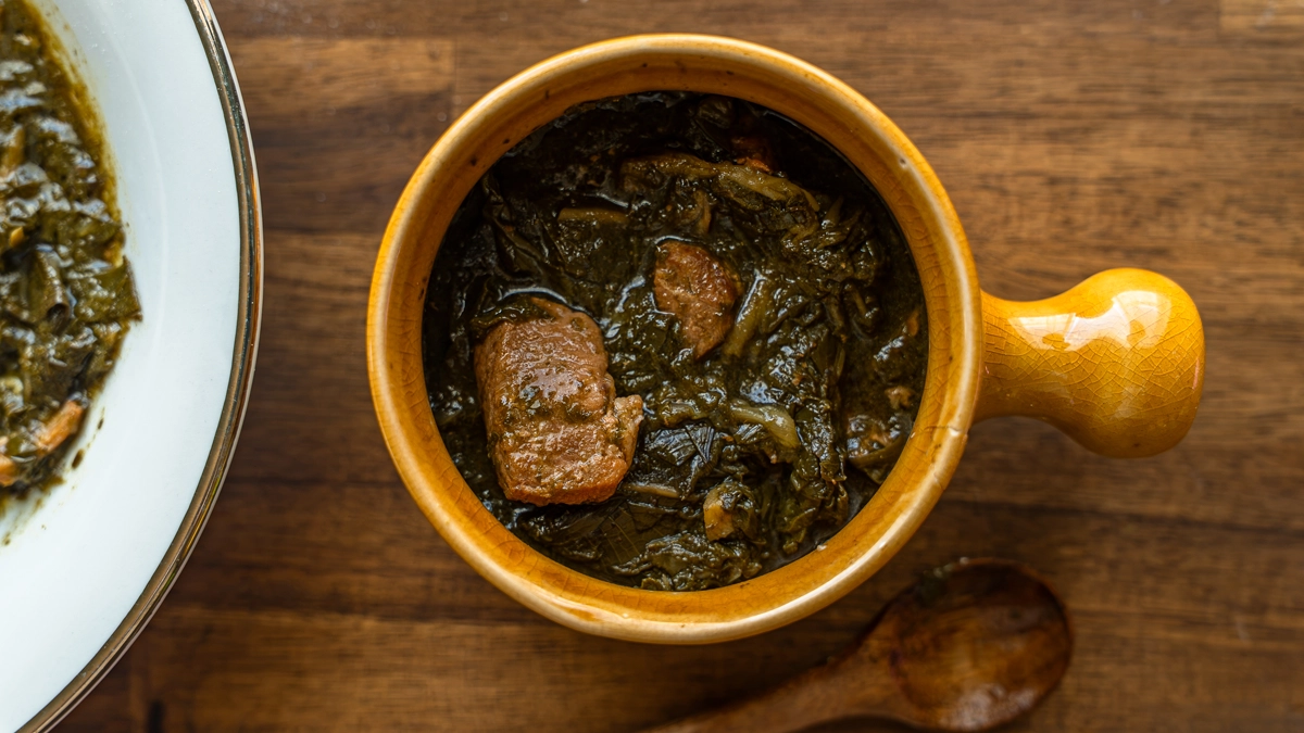 overhead close-up of luau stew in a small yellow ceramic crock with a wooden handle, large chunks of pork nestled in dark taro leaves, a wooden spoon and second bowl of stew alongside