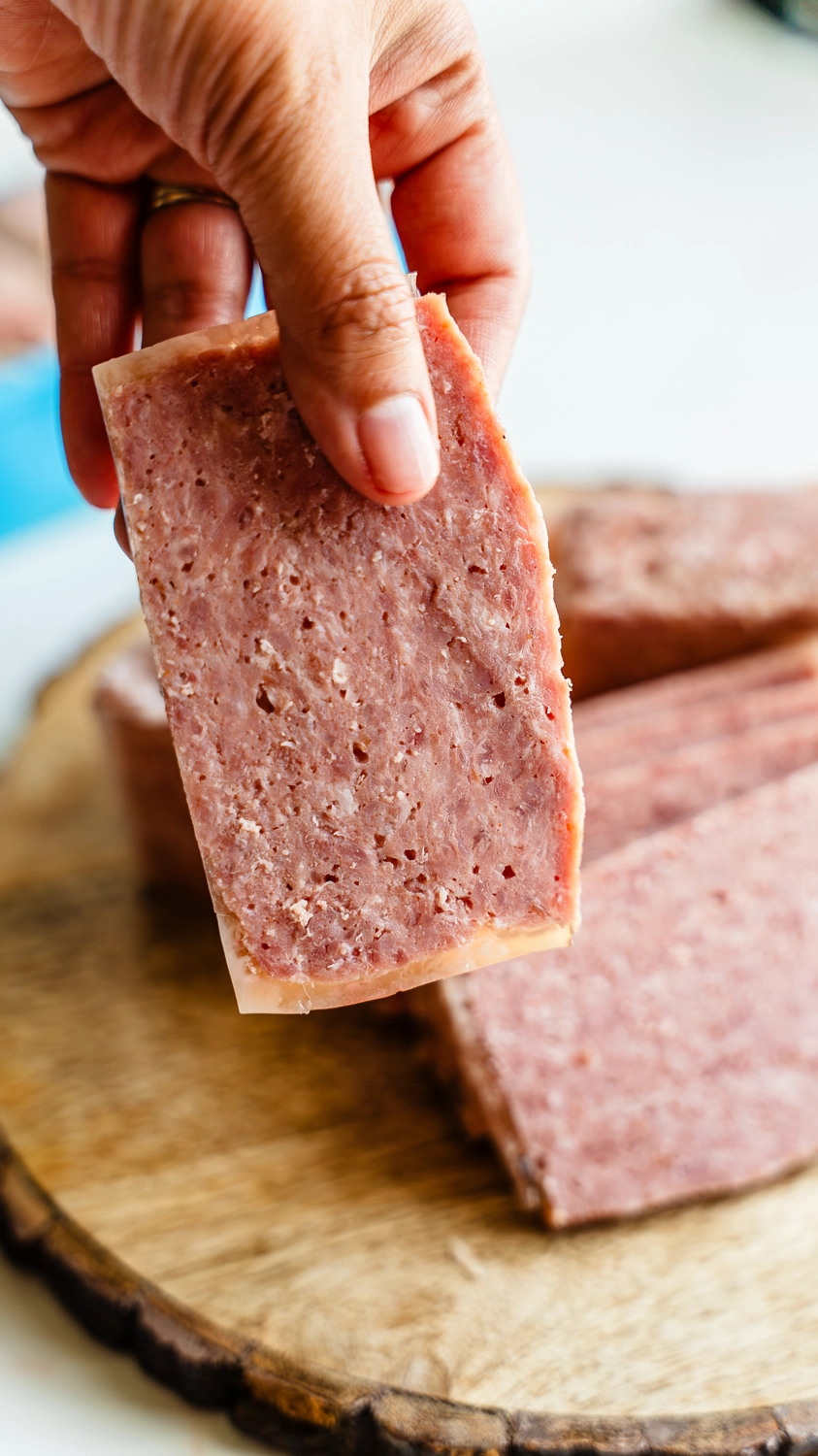 Hand holding up a thick slice of homemade SPAM showing the pink cured interior texture, with additional slices on a wood cutting board below
