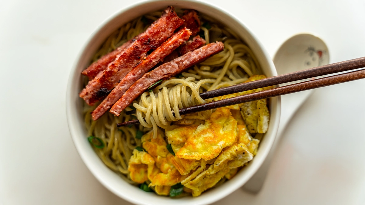 Overhead view of a bowl of saimin topped with crispy seared homemade SPAM strips, scrambled egg, and green onion, with chopsticks lifting noodles