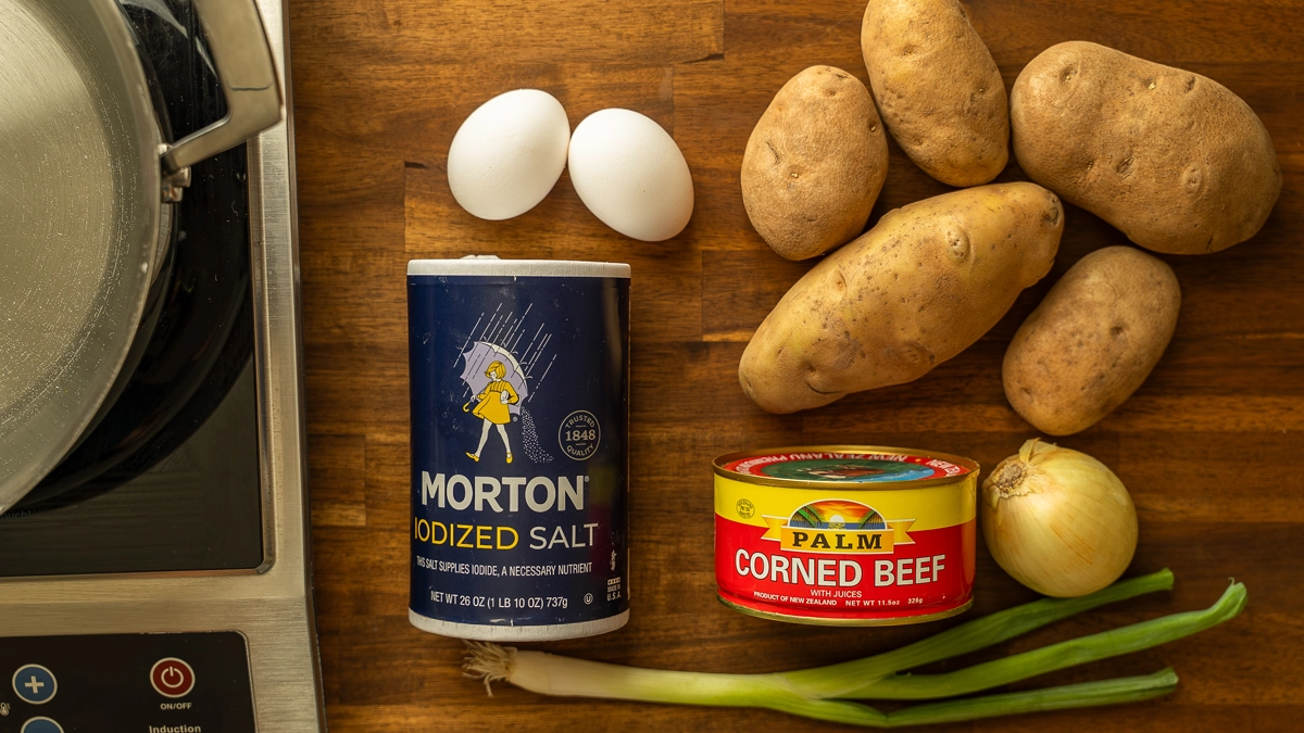 Ingredients for corned beef hash patties laid out on a wooden cutting board: eggs, russet potatoes, Morton salt, Palm corned beef, onion, and green onions
