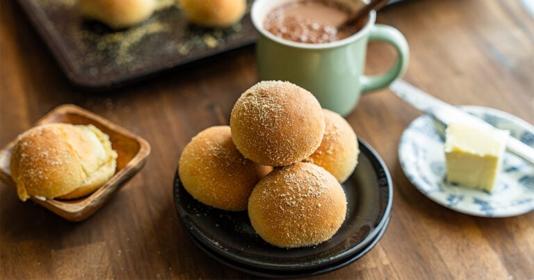 Stack of tangzhong pandesal rolls coated in fine breadcrumbs on a black plate, served with hot chocolate and a pat of butter.