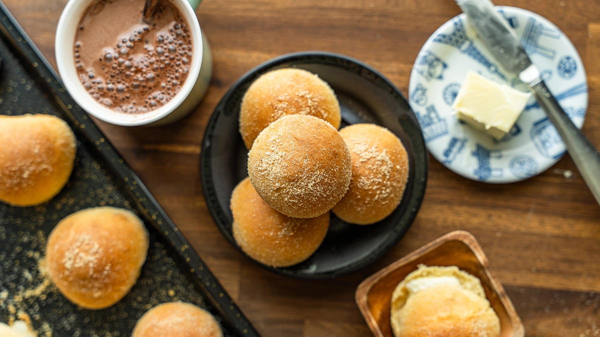 Overhead shot of baked pandesal rolls on a baking tray and plate, next to hot chocolate and a pat of butter.