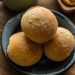 Close-up of soft pandesal rolls coated in fine breadcrumbs, stacked on a black plate.