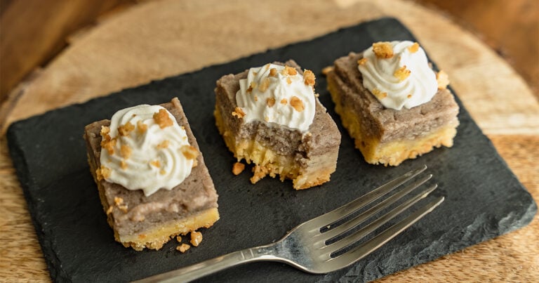 Overhead crop of kulolo crunch squares on a white tray and black slate board, topped with whipped cream and toasted macadamia crumble, styled with a striped napkin and dark plates on a wood table.